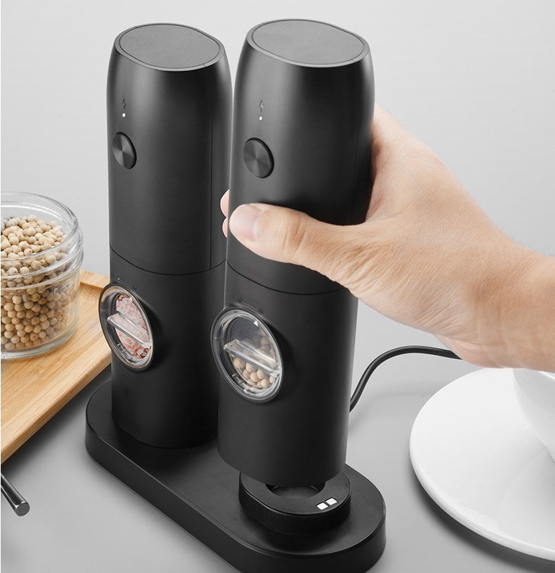 Black electric pepper grinder being held by a hand on a gray surface with a glass container of beans in the background.